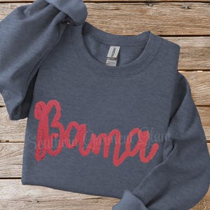 May include: A dark heather grey long-sleeve crewneck sweatshirt featuring the word Bama written in a textured red script font across the chest. The garment has a Gildan brand label at the inner neckline and long sleeves with ribbed cuffs.