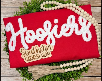 May include: A bright red t-shirt featuring the white chenille patch text "Hoosiers" in a cursive font. A wooden tag with the text "Southern GARMENT GLAM" is placed on the shirt, which is surrounded by a wooden bead garland.