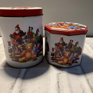 Op de afbeelding: Twee vintage kerstthema blikken met rode deksels. De grotere blik is cilindrisch en de kleinere blik is zeshoekig. Beide blikken hebben een witte achtergrond met een kleurrijke illustratie van Sinterklaas, een teddybeer en ander kerst speelgoed.