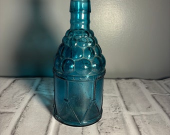 Peut inclure: Une bouteille en verre bleu avec un motif texturé en forme de raisin. La bouteille a un corps rond et un col étroit.