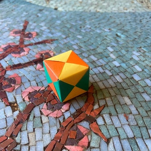 May include: A small origami cube made of paper with yellow, orange, and green squares. The cube is sitting on a mosaic tile surface.