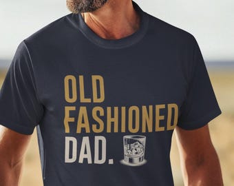 May include: A navy blue short-sleeved t-shirt featuring the gold and white text "OLD FASHIONED DAD." printed on the chest. Next to the word "DAD." is a small graphic illustration of a glass filled with an amber liquid and ice cubes.