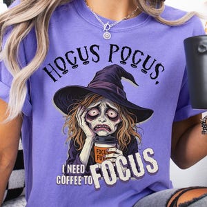 May include: A lilac-colored t-shirt with the phrase "Hocus Pocus" above a distressed witch graphic. The witch is holding a coffee cup that says "Focus Fuel". The text "I need coffee to focus" is below the graphic. The shirt is part of a Halloween-themed collection.