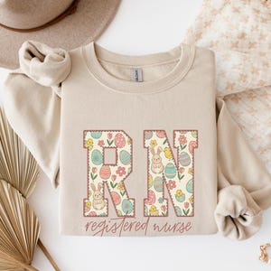 May include: Beige crewneck sweatshirt with the letters "RN" in a floral and Easter egg design. The words "registered nurse" are written in a cursive font below the letters. The sweatshirt has long sleeves and a relaxed fit.