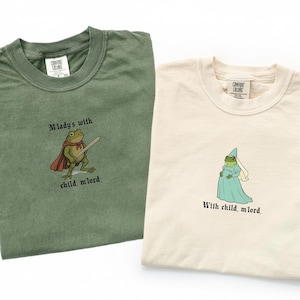 May include: Two Comfort Colors t-shirts, one olive green and one cream. The green shirt features a frog knight graphic with the text "M'lady's with child, m'lord." The cream shirt shows a pregnant frog in a gown, with the text "With child, m'lord."