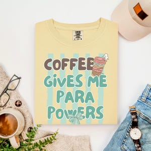 Puede incluir: Una sudadera Comfort Colors de color amarillo claro con el texto "COFFEE GIVES ME PARA POWERS" en marrón y verde azulado. Un gráfico de taza de café está encima de la palabra "coffee". La sudadera está rodeada por un sombrero, vaqueros, un reloj y una taza de café.