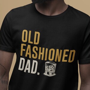 May include: A black short-sleeved t-shirt featuring the text "OLD FASHIONED DAD." in gold and cream lettering. Below the text is a graphic illustration of a glass filled with ice cubes, rendered in shades of white, gray, and black.