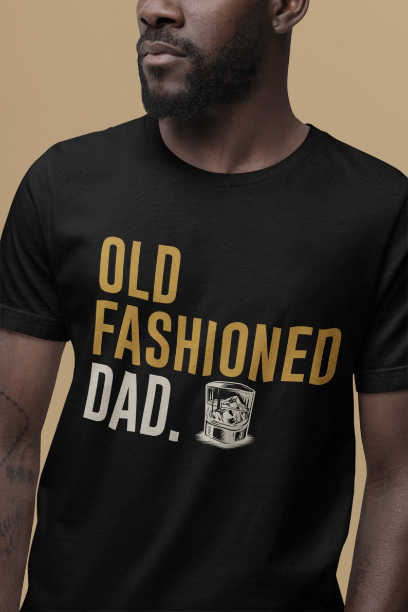 May include: A black short-sleeved t-shirt featuring the text "OLD FASHIONED DAD." in gold and cream lettering. Below the text is a graphic illustration of a glass filled with ice cubes, rendered in shades of white, gray, and black.