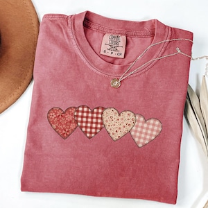 May include: A red-pink Comfort Colors t-shirt with four heart appliqués in floral and gingham patterns. A silver necklace with a pendant rests on the shirt. A brown hat and dried plant are in the background.