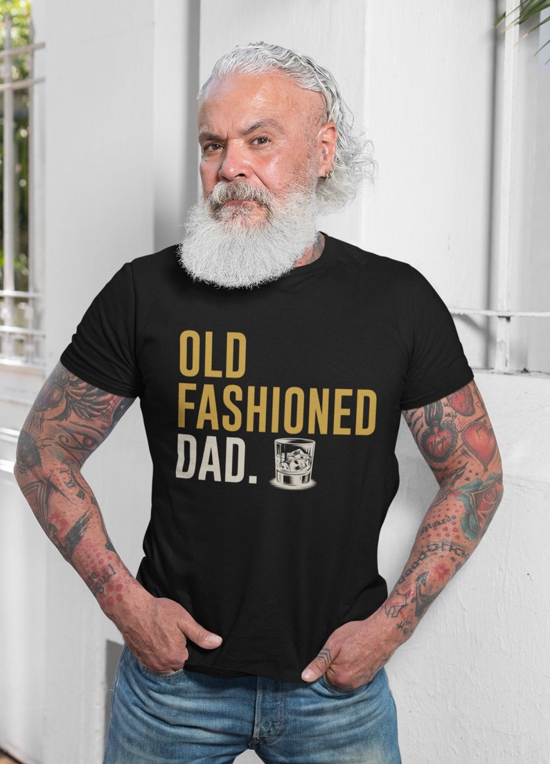 May include: A black short-sleeved t-shirt featuring the gold text OLD FASHIONED DAD and a graphic illustration of a glass containing a drink with ice cubes. The shirt is worn with blue denim jeans, showcasing a casual and standard fit design.