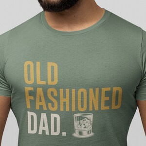 May include: A sage green short-sleeved t-shirt featuring the text "OLD FASHIONED DAD." printed in gold and white lettering. The design includes a graphic illustration of a glass filled with liquid and ice cubes next to the word DAD.