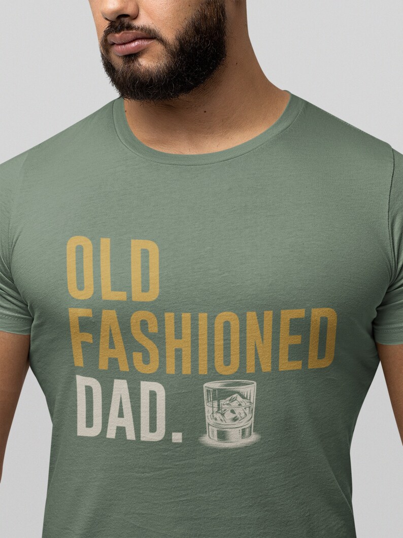 May include: A sage green short-sleeved t-shirt featuring the text "OLD FASHIONED DAD." printed in gold and white lettering. The design includes a graphic illustration of a glass filled with liquid and ice cubes next to the word DAD.