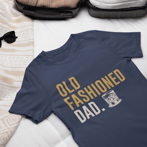May include: A navy blue short-sleeve t-shirt featuring the gold and white text "OLD FASHIONED DAD." printed on the front, accompanied by a graphic illustration of a glass containing a cocktail with ice cubes.