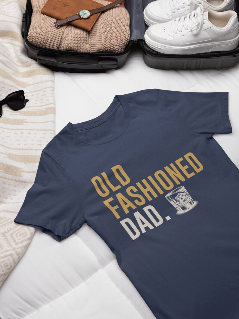 May include: A navy blue short-sleeve t-shirt featuring the gold and white text "OLD FASHIONED DAD." printed on the front, accompanied by a graphic illustration of a glass containing a cocktail with ice cubes.