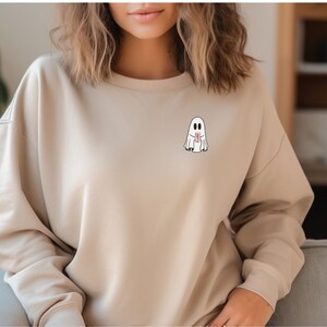 K&ouml;nnte beinhalten: Beiges Sweatshirt mit einem kleinen Cartoon-Geist-Motiv auf der Brust. Der Geist ist wei&szlig; mit schwarzen Augen und h&auml;lt einen rot-wei&szlig;en Gegenstand. Das Sweatshirt hat einen Rundhalsausschnitt und lange &Auml;rmel.