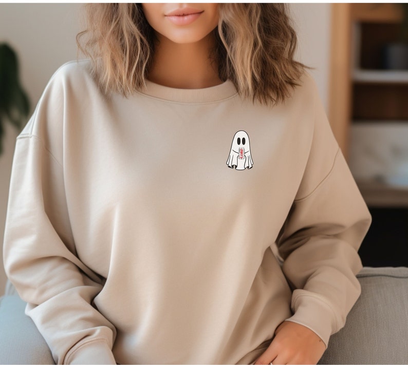 K&ouml;nnte beinhalten: Beiges Sweatshirt mit einem kleinen Cartoon-Geist-Motiv auf der Brust. Der Geist ist wei&szlig; mit schwarzen Augen und h&auml;lt einen rot-wei&szlig;en Gegenstand. Das Sweatshirt hat einen Rundhalsausschnitt und lange &Auml;rmel.