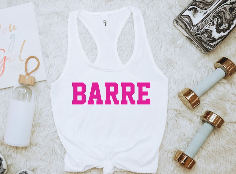 K&ouml;nnte beinhalten: Wei&szlig;es Tanktop mit dem Wort "BARRE" in leuchtend pinkfarbenen Blockbuchstaben. Das Tanktop ist unten geknotet. Zwei hellblaue Kurzhanteln mit goldenen Enden befinden sich im Hintergrund.