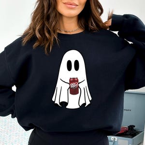 May include: Black sweatshirt with a white ghost design holding a Dr Pepper can. The ghost has black oval eyes and a simple, flowing design. The Dr Pepper can is red with white text.