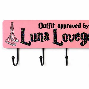 Puede incluir: Perchero de pared de madera rosa con el texto "Outfit approved by Luna Lovegood" y un conejo de dibujos animados con gafas. Incluye cinco ganchos negros para colgar objetos. Presenta un par de zapatillas rojas.