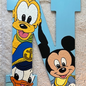 Peut inclure: Une lettre N en bois bleu clair ornée d'illustrations peintes à la main de Pluto, Donald Duck portant un chapeau de marin bleu avec un emblème d'ancre dorée, et Mickey Mouse en chemise bleue. La surface présente une texture de pinceau visible.