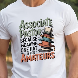 May include: White t-shirt featuring the text "Associate Pastor" in green and orange, with a stack of colorful hats. The text continues, "BECAUSE WEARING ONE HAT IS FOR AMATEURS."