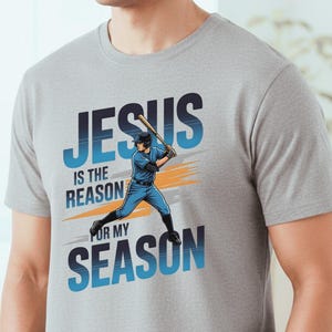 May include: A light gray t-shirt featuring the text "JESUS is the REASON FOR MY SEASON" in blue and white. A baseball player in blue uniform is depicted swinging a bat. The shirt is a crew neck style.