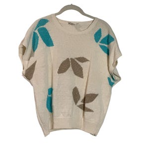 May include: A white knitted sweater with short sleeves and a crew neck. The sweater has a leaf pattern in shades of brown and teal.