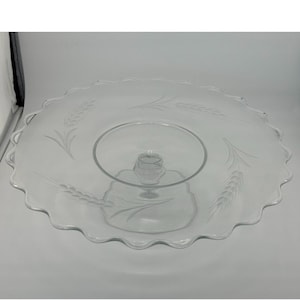 May include: A clear glass cake stand with a scalloped edge and a pedestal base. The stand is decorated with an etched wheat design.
