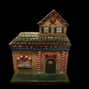May include: A decorative miniature toy store with a glittery green roof, red and white striped columns, and a green base. The building has a red heart on the roof and the word "TOYS" above the door. The window has the word "Treats".