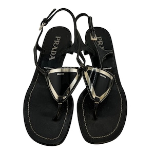 May include: A pair of black leather thong sandals featuring a silver-tone triangular plaque with the Prada logo on the front. The sandals have a slingback strap with a metal buckle, white contrast stitching along the edges, and a low block heel.