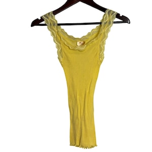 May include: A bright yellow ribbed tank top featuring a scoop neckline and wide shoulder straps adorned with delicate scalloped lace trim. The garment is displayed on a dark brown hanger and has a straight hemline with a subtle ruffled edge.