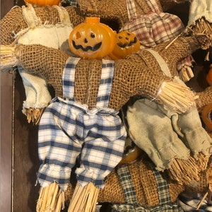 May include: A collection of scarecrow dolls featuring orange plastic jack-o'-lantern heads and bodies made of brown burlap fabric. The dolls wear overalls in various plaid patterns of blue, white, and green, with straw-like material at the ends of limbs.