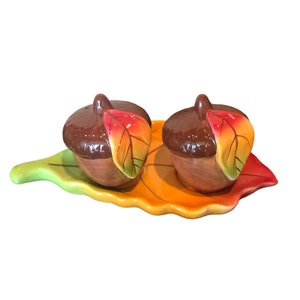 May include: A ceramic salt and pepper shaker set featuring two acorn-shaped shakers with brown tops and tan bases, each with a colorful leaf accent. The shakers sit on a leaf-shaped tray with green, yellow, and orange hues, perfect for fall decor.