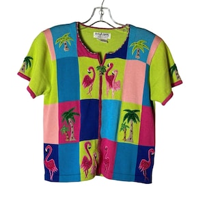 Puede incluir: Un cárdigan de manga corta multicolor con cierre de cremallera. El cárdigan presenta un diseño de patchwork con cuadrados rosas, azules, verdes y blancos. Cada cuadrado tiene imágenes bordadas de flamencos rosas y palmeras. El cárdigan tiene el texto "JackJill Queen" en la etiqueta.