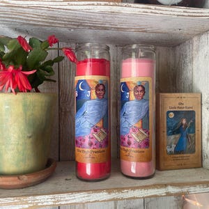 Könnte beinhalten: Zwei Glas-Kerzen, eine rote und eine rosa, mit Etiketten mit einer Illustration einer Person und den Worten "The High Priestess". Die Kerzen stehen auf einem Holzregal mit einer kleinen Topfpflanze und einer Tarotkarte.