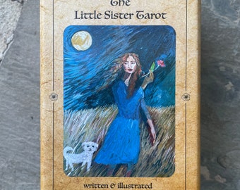 May include: The box for The Little Sister Tarot deck features a vintage-style design with a brown background and gold accents. The title "The Little Sister Tarot" is printed in black lettering. The front of the box shows an illustration of a woman in a blue dress holding a flower, with a white dog at her feet. The text "written & illustrated by Ginny Thonson" is printed below the illustration.