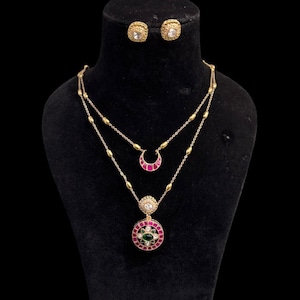 Könnte beinhalten: Ein goldfarbenes Schmuckset mit Halskette und Ohrringen. Die Halskette hat zwei Stränge, einen mit einem halbmondförmigen Anhänger und einen mit einem kreisförmigen Anhänger mit roten und grünen Akzenten. Die Ohrringe sind quadratisch.