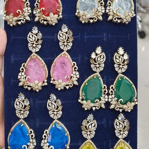 Peut inclure: Une collection de boucles d'oreilles en forme de goutte d'eau avec des pierres précieuses de différentes couleurs, dont rouge, transparent, rose, vert, bleu et jaune. Chaque boucle d'oreille présente un motif filigrané doré avec des accents de pierres transparentes.