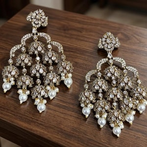 Può includere: Un paio di orecchini a lampadario decorati con molteplici pendenti a forma di campana incastonati con cristalli trasparenti e accentuati da piccole perle bianche. La montatura è color oro e il design include un perno a forma di fiore.