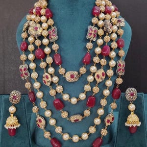 May include: A multi-strand necklace and matching earrings set. The necklace features alternating strands of pearls, red gemstones, and gold-toned decorative elements. The earrings have a similar design, with a red gemstone and gold accents.