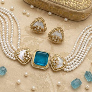 Puede incluir: Un collar de gargantilla de perlas de varias hebras con una piedra preciosa azul rectangular central, flanqueada por dos acentos triangulares de nácar blanco con filigrana dorada y pequeñas piedras claras, junto con pendientes a juego.