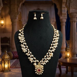 May include: A pearl necklace and matching earrings set displayed on a black mannequin. The necklace features multiple strands of white pearls with a gold pendant and a red gemstone. The earrings are small pearl clusters.