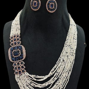 May include: A necklace and earring set featuring a multi-strand beaded necklace with a large square pendant and matching earrings. The pendant and earrings have a dark blue center stone surrounded by smaller blue stones and a rose gold-colored frame.