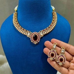 Può includere: Un set di collana e orecchini color oro. La collana e gli orecchini presentano una pietra centrale ovale, rosso scuro, circondata da pietre rosse e chiare più piccole. I gioielli sono esposti su un manichino blu.