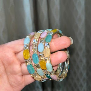 Puede incluir: Un conjunto de cuatro pulseras coloridas. Cada pulsera está adornada con una variedad de piedras preciosas ovaladas multicolores, incluyendo amarillo, azul, verde y rosa. Cristales transparentes de corte cuadrado se intercalan entre las piedras preciosas.