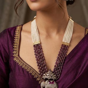 Puede incluir: Collar de múltiples hilos con perlas blancas y cuentas rojas oscuras. El colgante presenta un diseño de elefante con detalles intrincados y un paraguas enjoyado. Pendientes a juego, todo sobre una blusa morada.