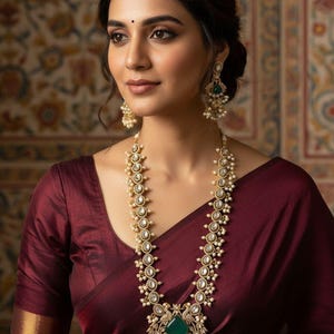 Puede incluir: Una mujer con un sari de seda granate y joyas de oro. Las joyas incluyen un collar largo con perlas y piedras transparentes, y pendientes a juego. El collar tiene un gran colgante de piedra verde.