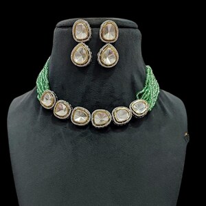 May include: A necklace and earring set displayed on a black mannequin. The necklace features large, clear, teardrop-shaped stones with gold and silver accents, connected by a strand of green beads. The matching earrings have similar stones.