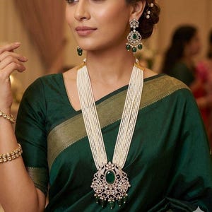 May include: A woman wearing a dark green sari and a multi-strand pearl necklace with a large pendant. The pendant features a large oval green stone surrounded by smaller stones. Matching earrings with green stones complete the look. A gold bracelet is visible on the left wrist.