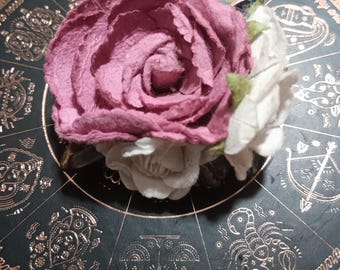 May include: A decorative accessory featuring a large, textured mauve paper rose layered with smaller white paper flowers and small green felt leaves, resting on a dark circular base with gold zodiac symbols and text.